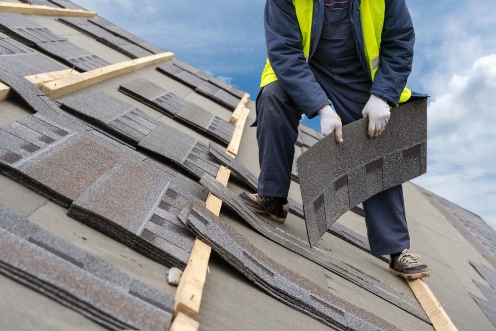 A worker in uniform holds and lays asphalt shingles on the roof of a house under construction