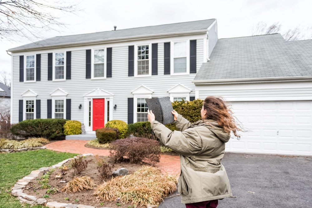 A homeowner standing in front of a house on a windy day, holding shingles and inspecting the damage