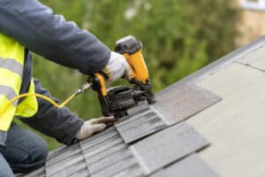 Unrecognizable roofer worker in uniform workwear using air or pneumatic nail gun and installing asphalt tiles