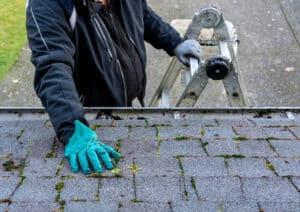 A man at the top of a ladder clears moss and dirt from the gutter of a house