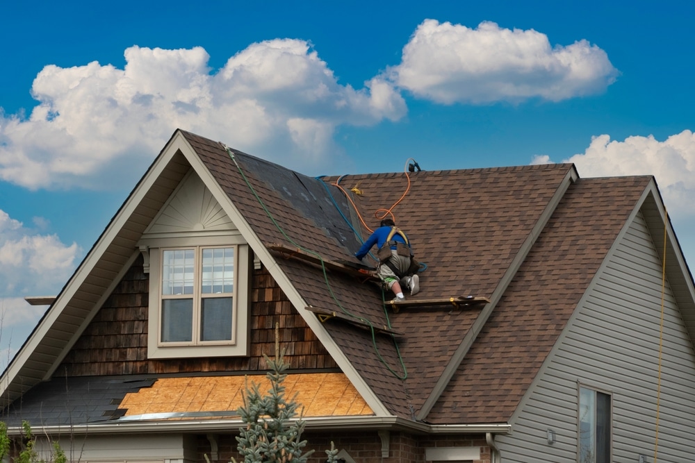 A male person kneeling on a sloped roof doing DIY roof repair for his house with grey roofing materials
