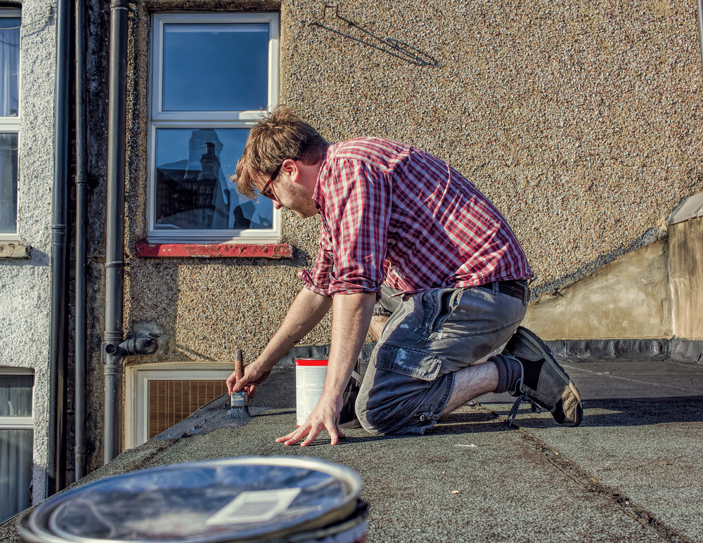 Young male homeowner in red plaid shirt sealing roof of his house, doing DIY, roof maintenance improvement