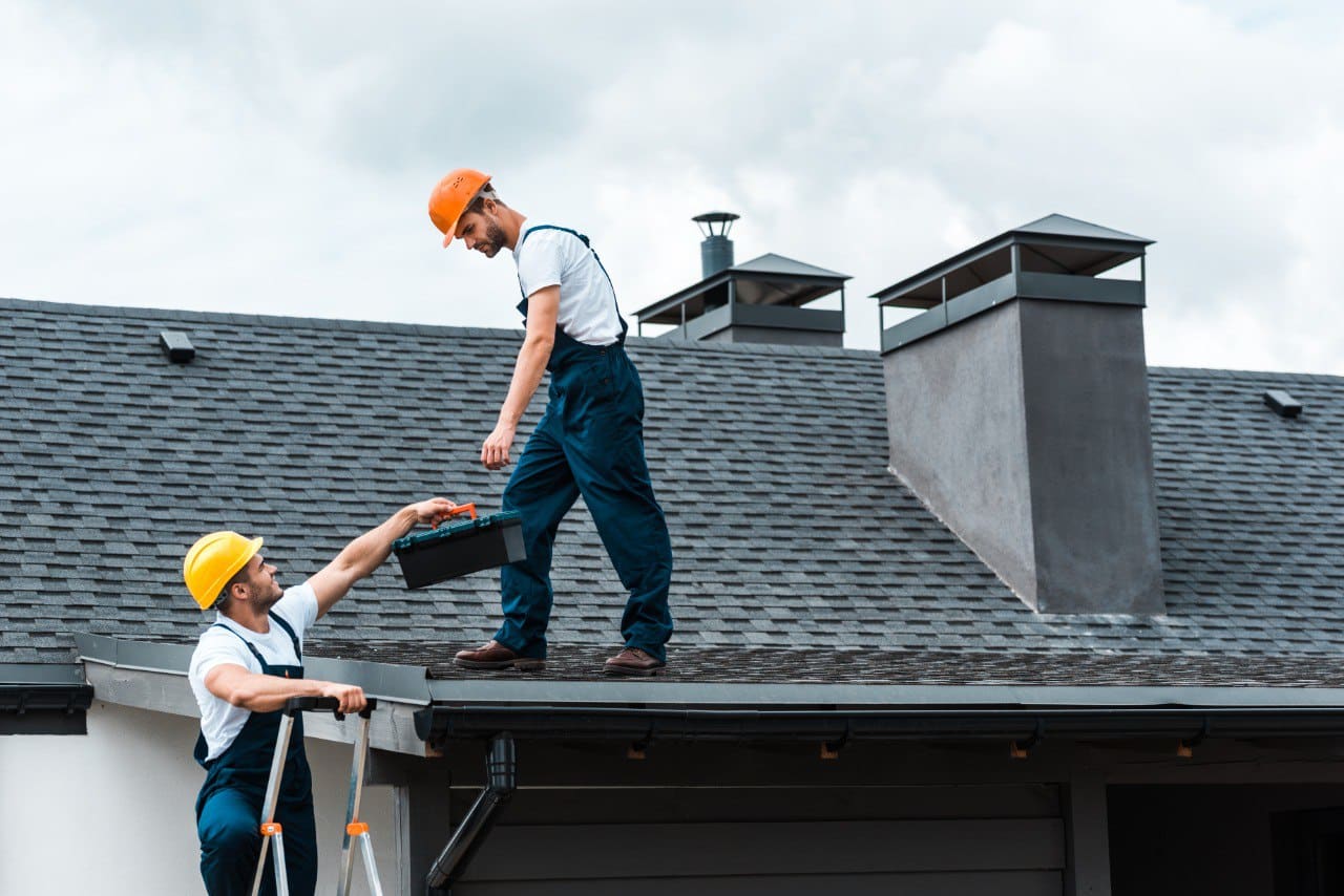 Handyman in helmet and blue uniform, giving toolbox to colleague, professional roofing expert standing on roofto