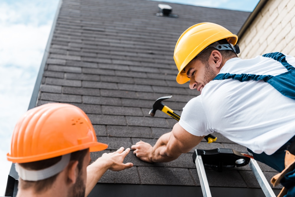 Low-angle view of a roof repairman holding a hammer while looking at a coworker.
