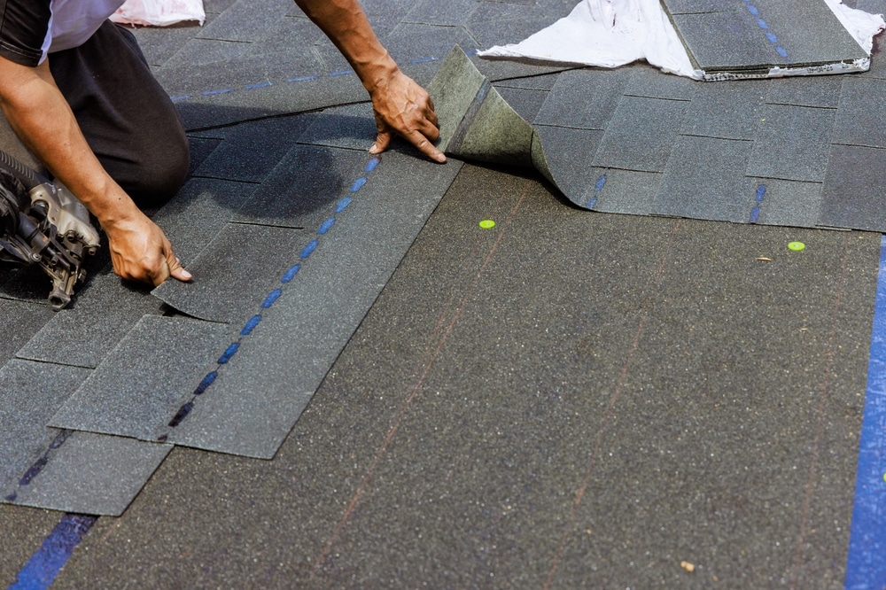 A construction worker installs new asphalt shingles on a house roof using a pneumatic nail gun