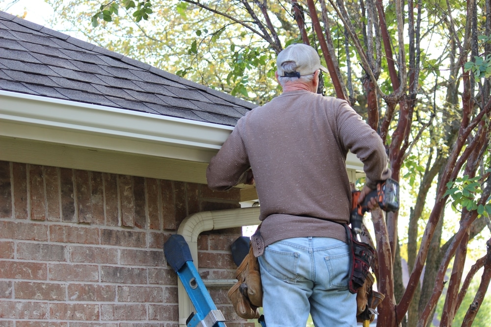 Man with equipment doing DIY, installing roof gutters on residential property, attaching aluminum rain gutter and down spout
