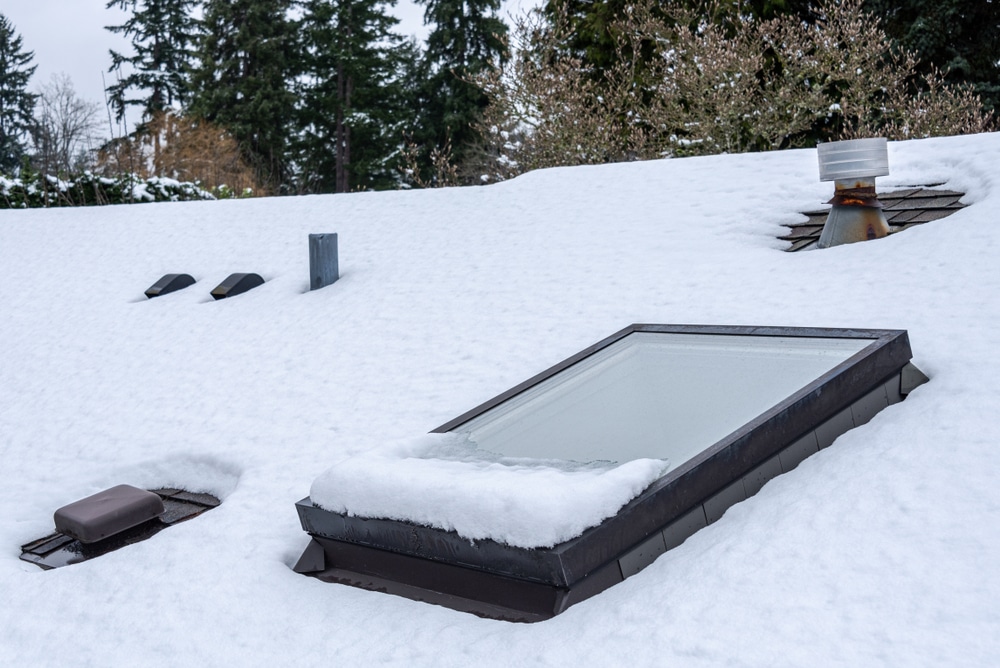 Residential house rooftop covered in fresh snow, skylight, roof vents, evergreen trees, skylight covered with snow