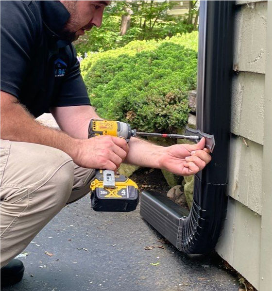 Technician securing a downspout to the exterior wall with a power drill during a gutter repair service in Boston MA