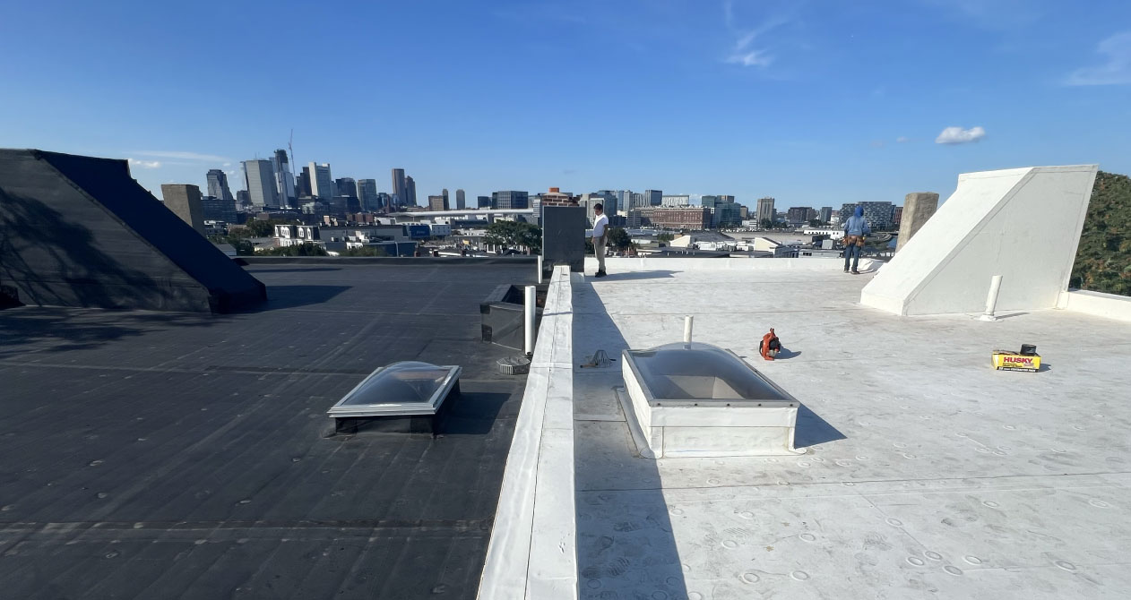 A rubber roofing in Boston MA with black and white sections, skylights, and chimneys inspecting the rooftop with city views