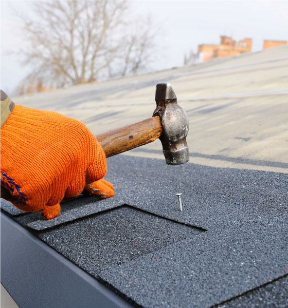 A worker wearing an orange glove uses a hammer to secure a nail into asphalt shingles during a roof repair in Boston