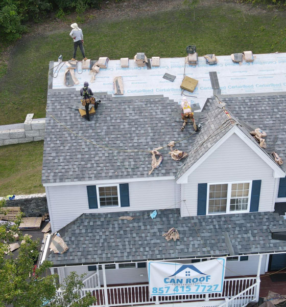 Workers are installing new shingles on a house during a roof replacement in Boston, with a Can Roof Construction banner displayed