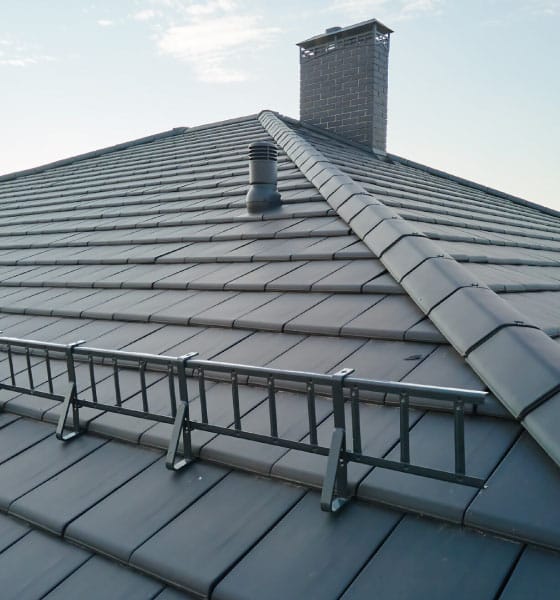 A slate roofing system with dark grey tiles, a chimney, ventilation pipes, and a mounted snow guard for safety