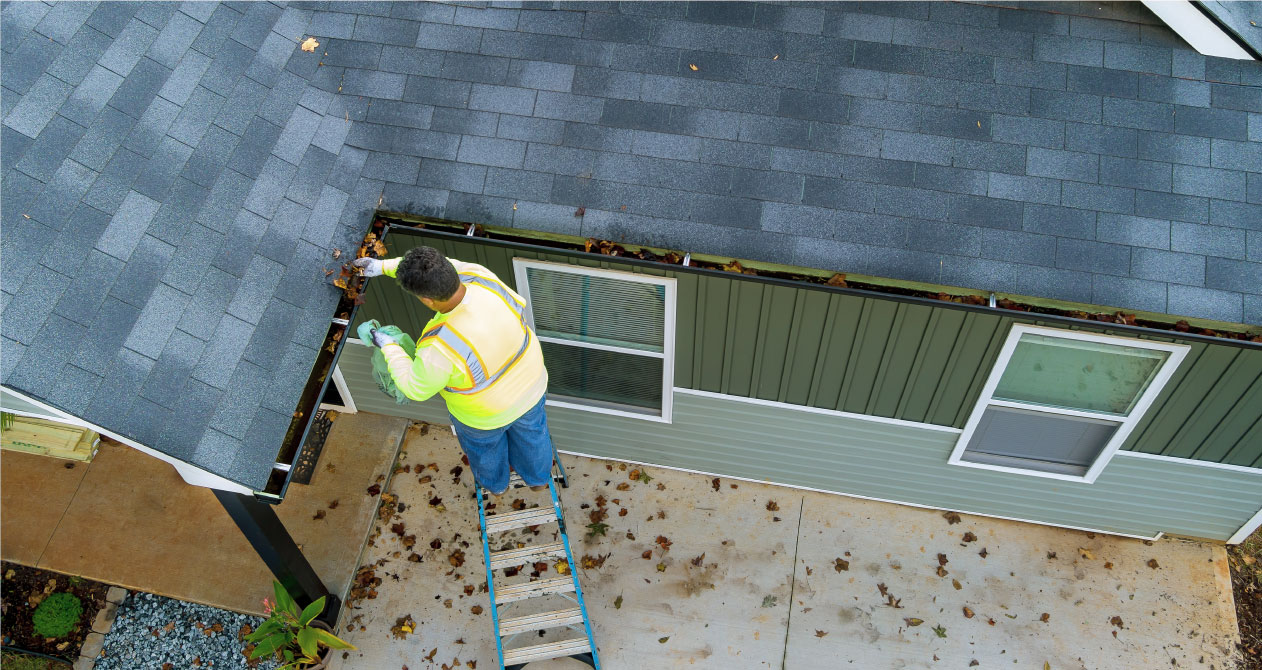 A worker in a vest stands on a ladder, removing leaves from a house’s gutter, performing gutter cleaning in Boston MA