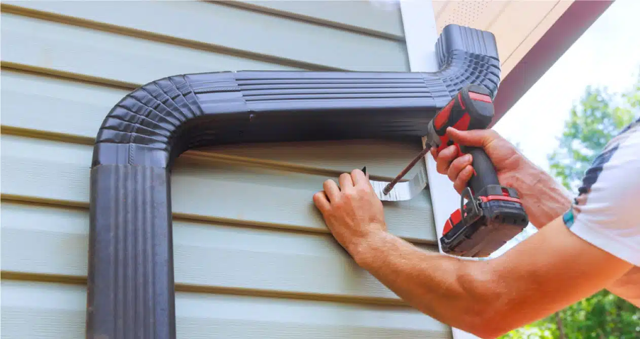 Gutter repair in Boston MA, a technician is securing a black downspout to the house using a power drill for water drainage