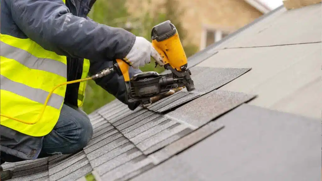 Roof contractor working in special protective work wear and gloves, using air or pneumatic nail gun and installing shingles