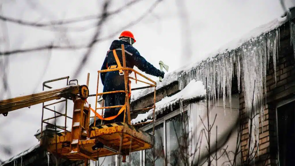 Utility worker in a safety harness and red helmet removing large icicles from a snow-covered roof using a lift in winter