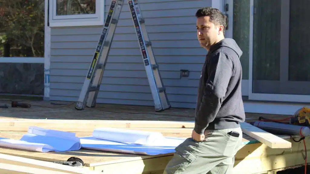 CAN Roof Construction worker in a dark hoodie and green pants standing on a partially built wooden deck.