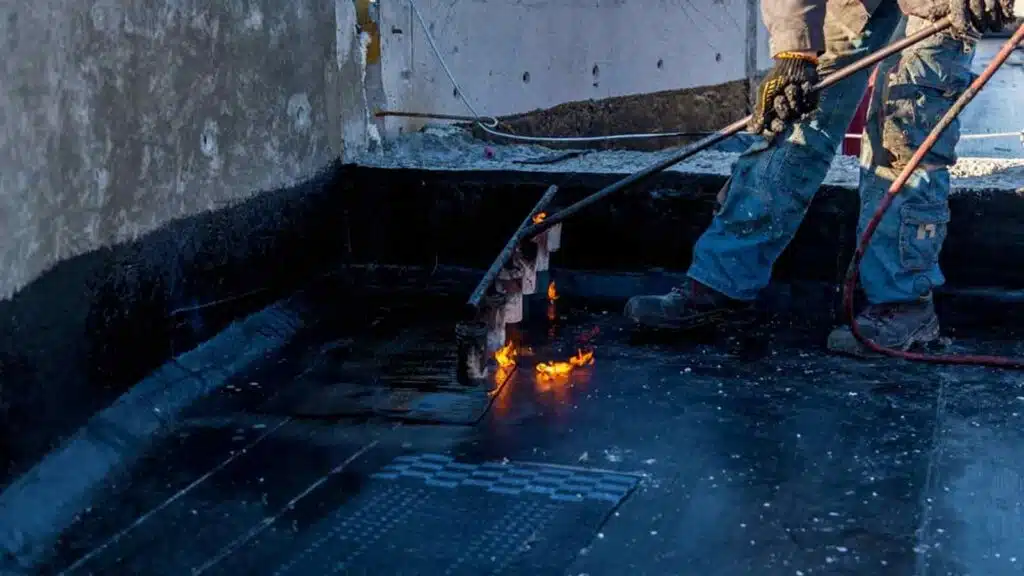 Worker installing a bituminous waterproofing membrane on a flat roof using a torch for secure sealing.