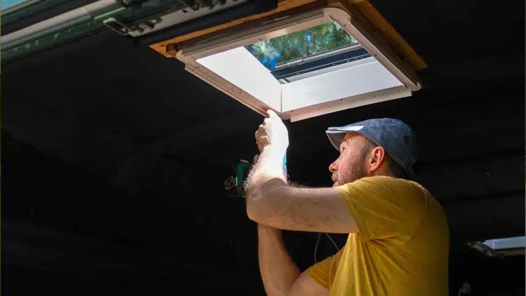 Handyman in yellow shirt installing camper van roof window with cordless drill under open skylight.