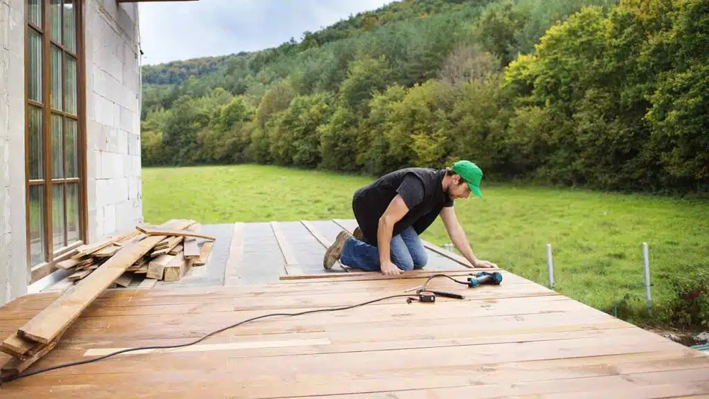 Handyman wearing a green cap and black shirt installing wooden flooring on an outdoor deck.