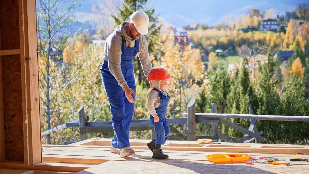 Father and toddler son wearing hard hats and overalls building a wooden frame house together.