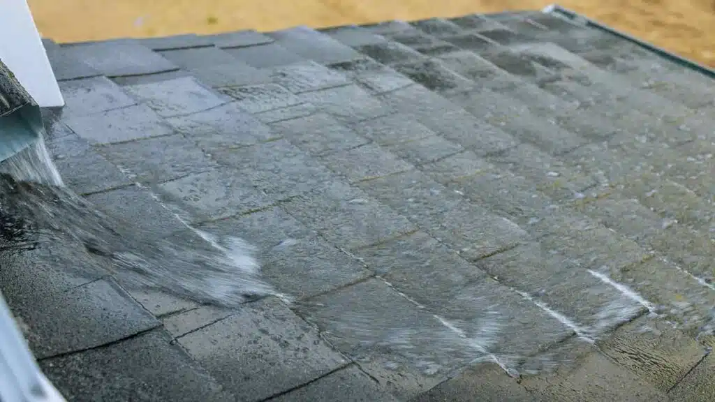 Heavy rainwater flowing down a dark asphalt shingle roof during a storm, with water draining off the edge.