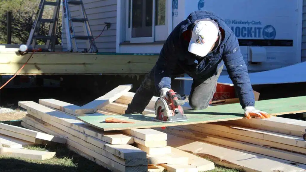 CAN Roof Construction worker using a circular saw to cut a wooden board on a construction site with stacked lumber.