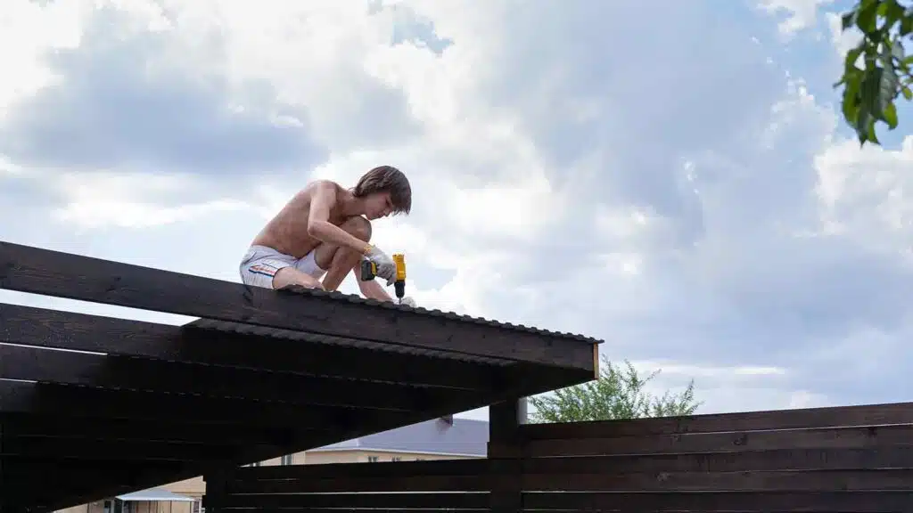 Young man repairing a metal roof while using a screwdriver, working at height under a partly cloudy sky.