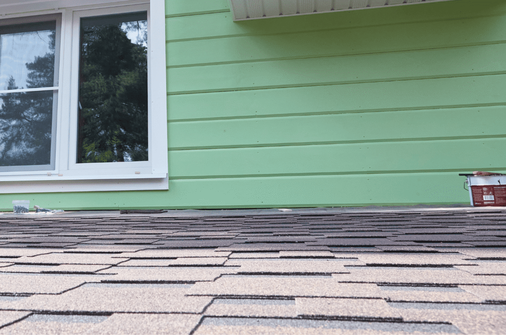 Close-up of asphalt roof shingles beside a house wall and window, showing a residential roof repair area