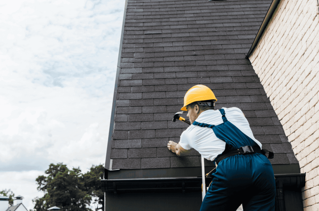 Professional roofer with a hard hat using a hammer to repair shingles on a residential roof after a temporary roof leak