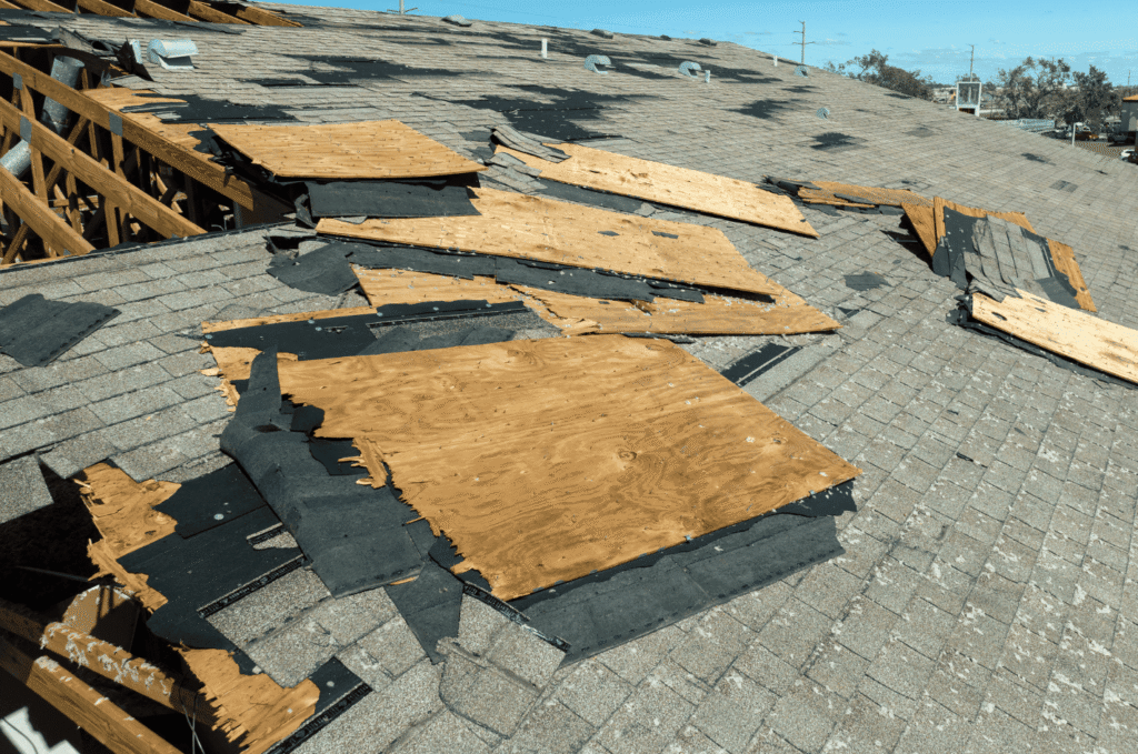 Severe storm damage roof with torn shingles, exposed plywood roof decking, and structural framing visible after high winds