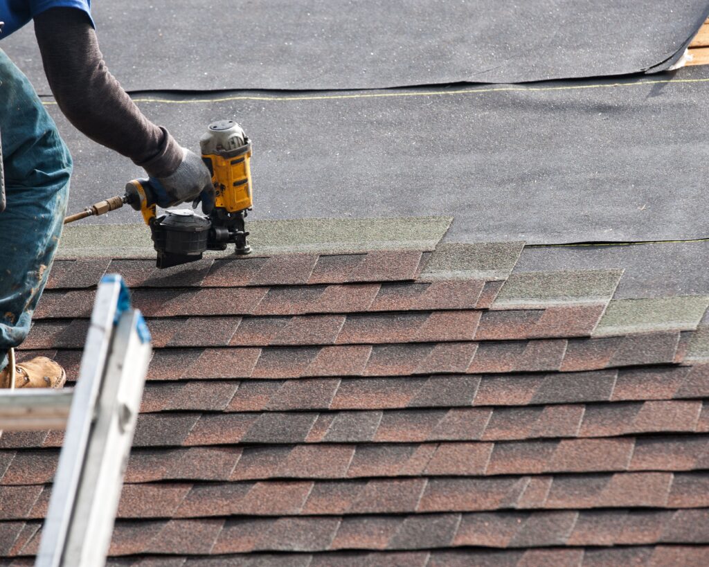 Roofer nailing architectural shingles with a pneumatic gun over roof underlayment from a ladder.