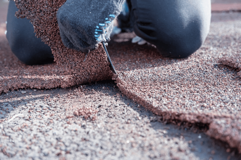 Close-up view of a professional roofer wearing gloves and cutting damaged roofing shingles with a knife during roof repair