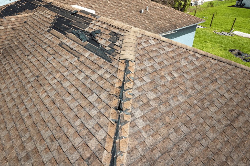 Close-up of storm-damaged roof showing lifted ridge cap shingles and missing asphalt shingles with exposed underlayment