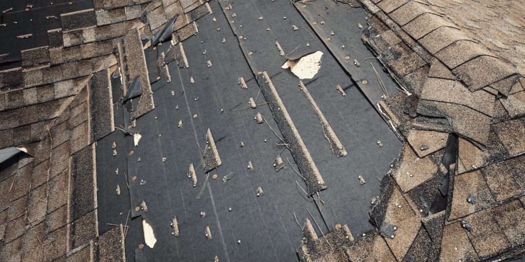Close-up of severe roof damage showing missing asphalt shingles, exposed black underlayment, and scattered roofing nails