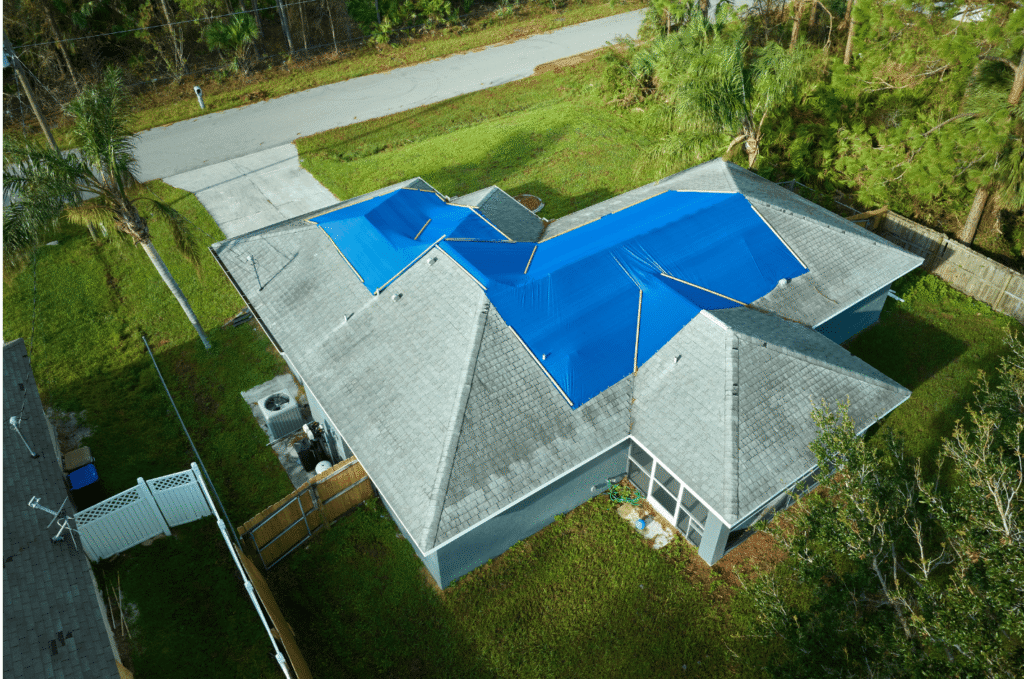 Storm-damaged residential roof covered with blue emergency tarps secured with lumber strips and scattered shingle debris