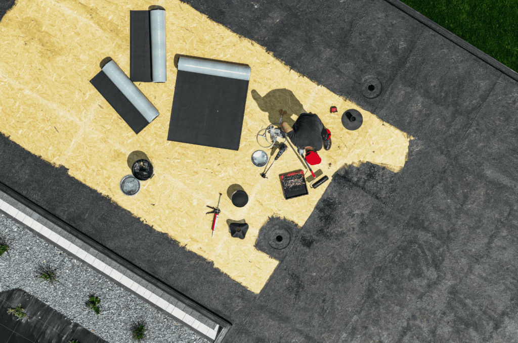 Flat roof worker applying membrane repair, showing surface wear and drainage areas that should be checked after heavy rain.